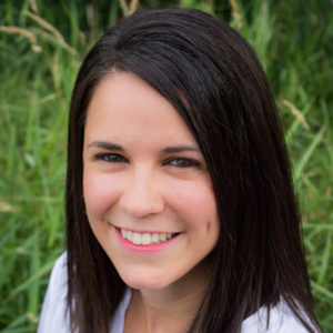 Latinx woman with dark brown hair and brown eyes smiling with blurred outdoor background